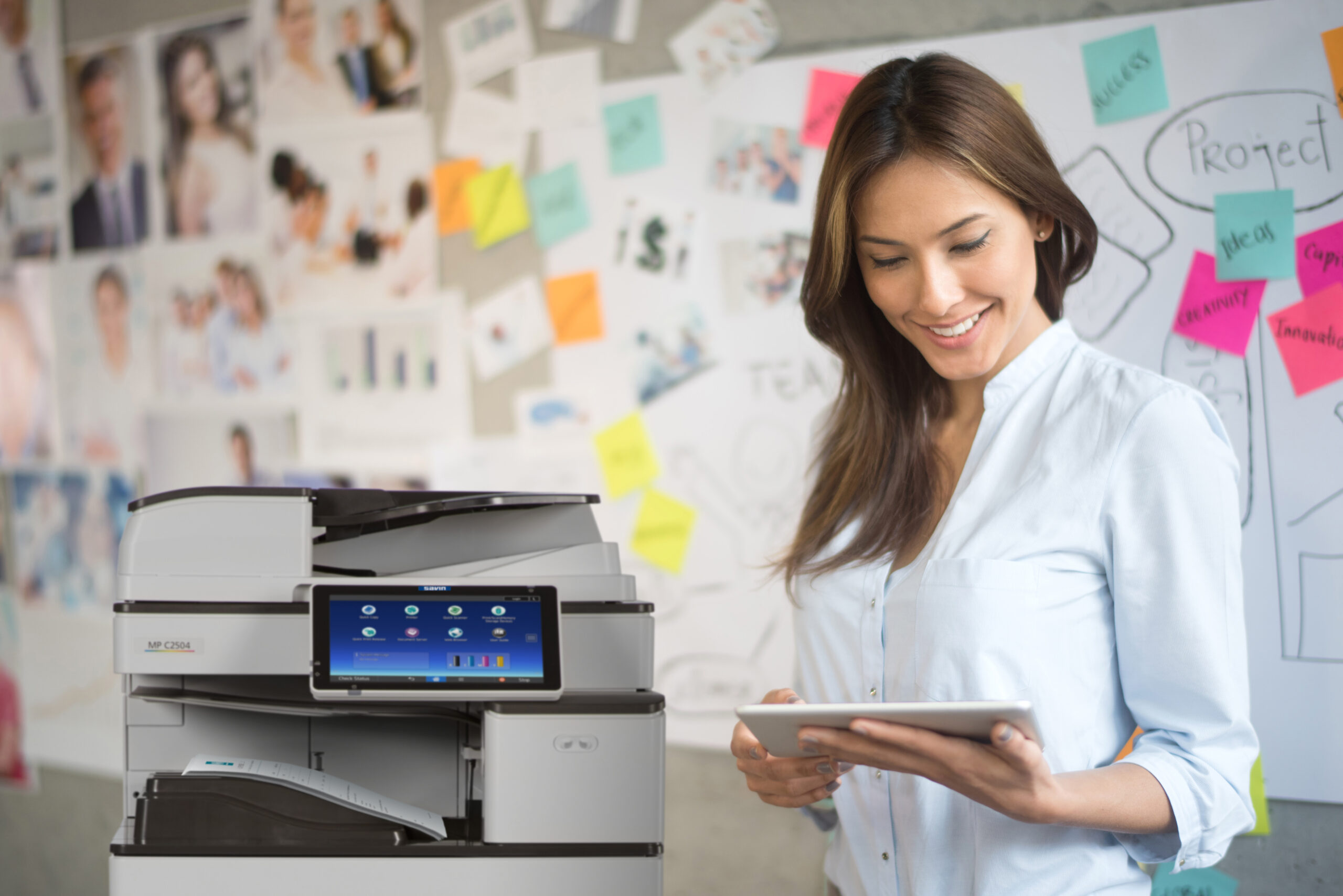 A smiling Asian woman using a tablet standing in front of a Ricoh Multifunctional device in a modern office environment.