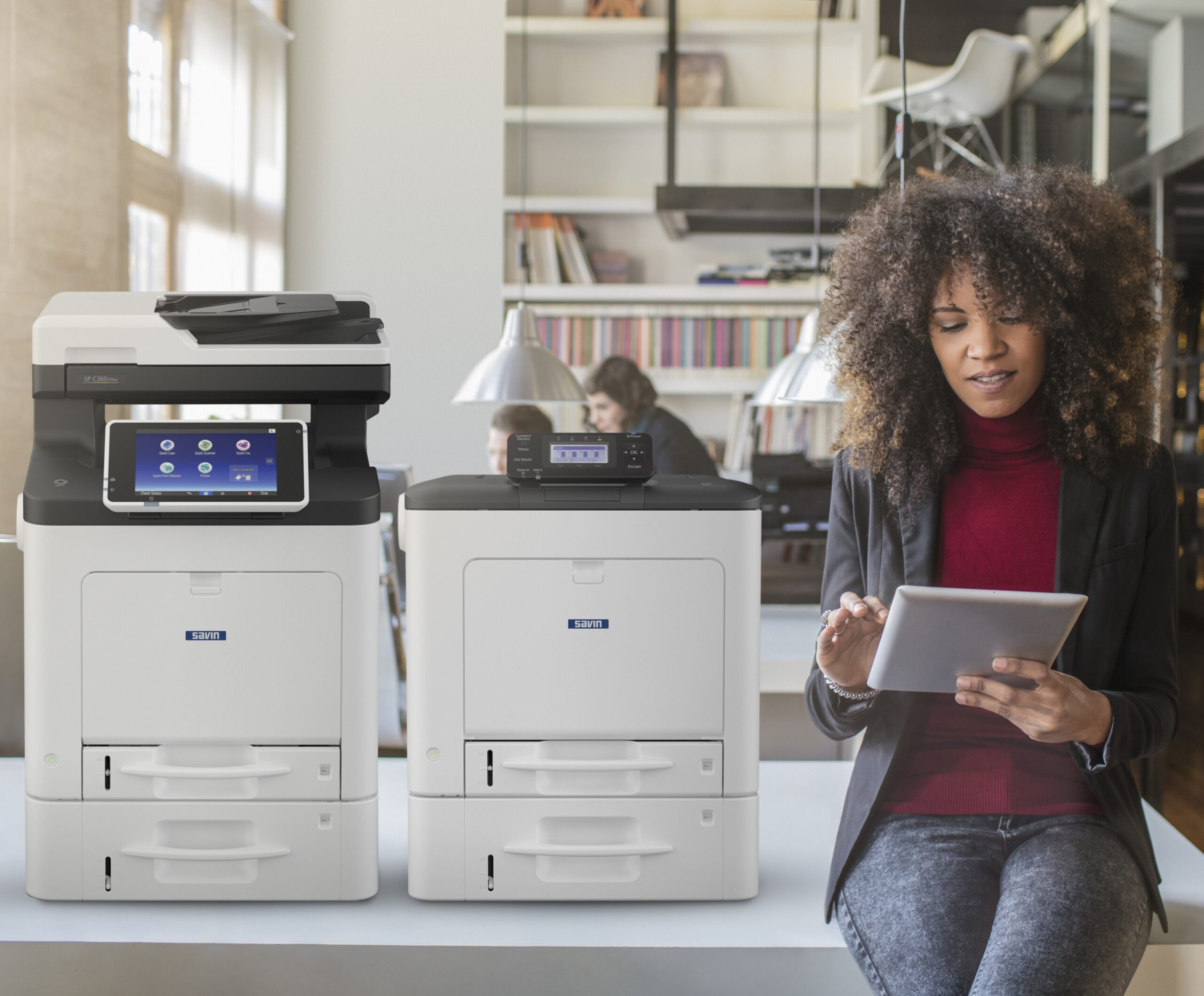 A multitasking Black woman effortlessly manages her work with a tablet while two Ricoh/Savin multifunctional devices printers hum in the background. Stay productive with our Business Technology Solutions like Commercial Printers, Commercial Copiers, and other Multifunctional Devices