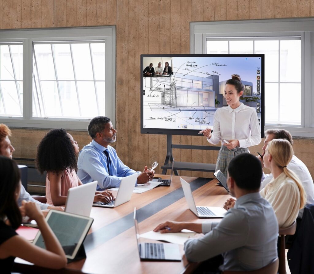 A young Asian woman—an architectural leader, leading a meeting using a large Ricoh Interactive Whiteboard in place of a traditional whiteboard. The screen displays illustrated visual aids rich in color and design, in front of a conference room of engaged professionals sitting at a long rectangular conference table, all taking notes on laptops.